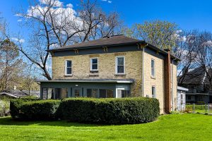 The front of a brick house with hedges in front and a grassy lawn on a sunny, early spring day