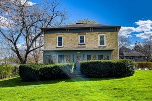 The front of a brick house with hedges in front and a grassy lawn on a sunny, early spring day